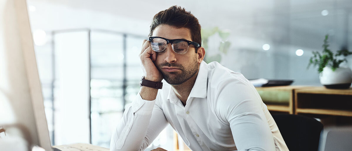 Müder Mann sitzt an seinem Schreibtisch, hat die Augen geschlossen und seinen Kopf auf einen Arm aufgestützt.