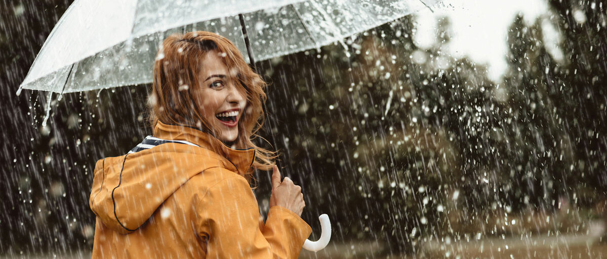 Eine Frau läuft lachend mit einem durchsichtigen Regenschirm und Regenjacke durch den Regen.