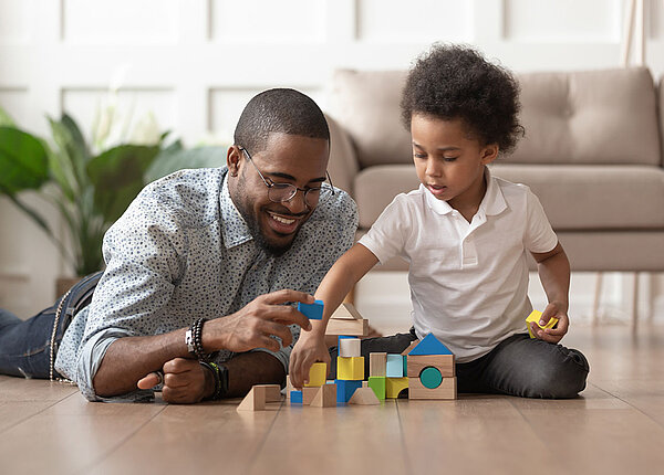 Vater und Sohn (beide afro-amerikanisch) spielen mit Holzspielzeug auf dem Boden eines Wohnzimmers.