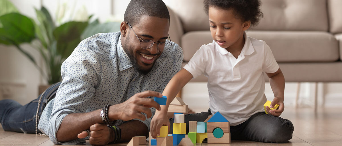 Vater und Sohn (beide afro-amerikanisch) spielen mit Holzspielzeug auf dem Boden eines Wohnzimmers.