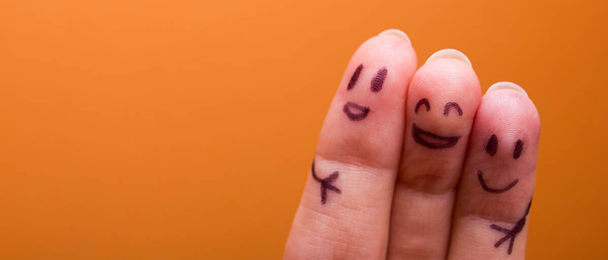 Drei Finger sind mit Gesichtern bemalt. Der mittlere hat die Arme um die beiden anderen geschlungen. Alle drei lachen, der mittlere am stärksten.