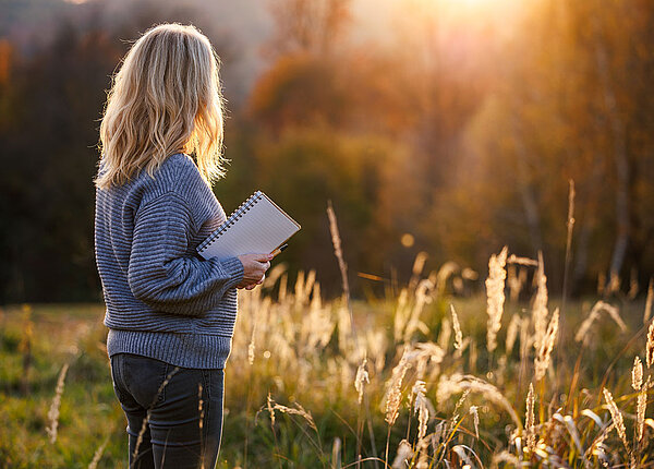Rückansicht einer blondhaarigen Frau, die in der rechten Hand einen Block und Stift hält und sich den Sonnenuntergang anschaut.