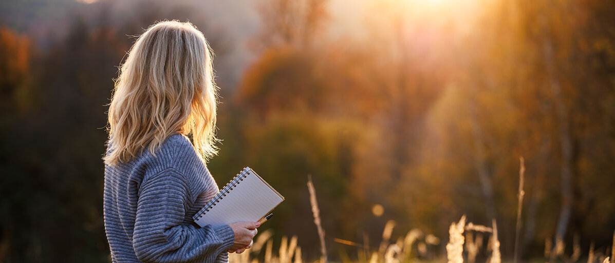 Rückansicht einer blondhaarigen Frau, die in der rechten Hand einen Block und Stift hält und sich den Sonnenuntergang anschaut.