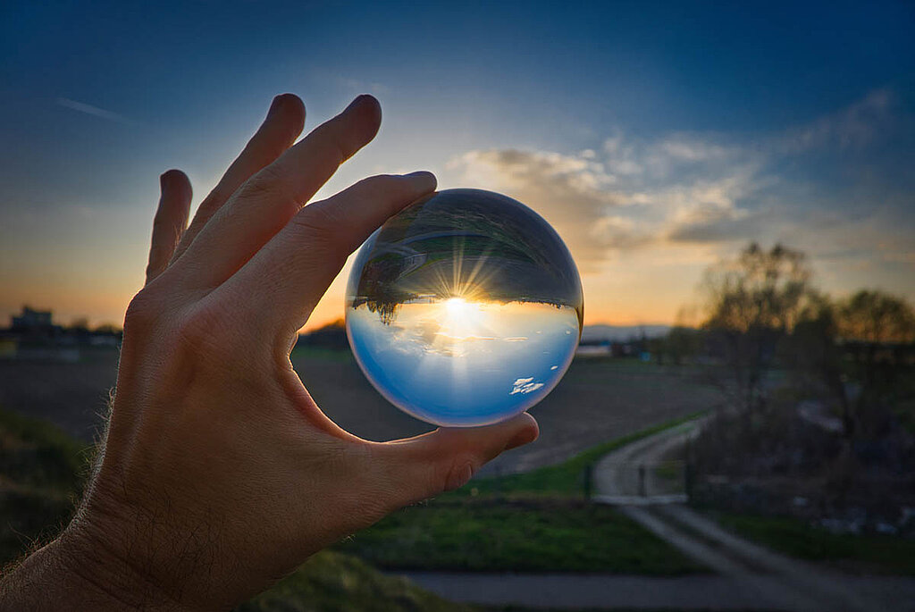 Eine Hand hält eine Kristallkugel. Wir blicken durch die Kugel hindurch in die untergehende Sonne.