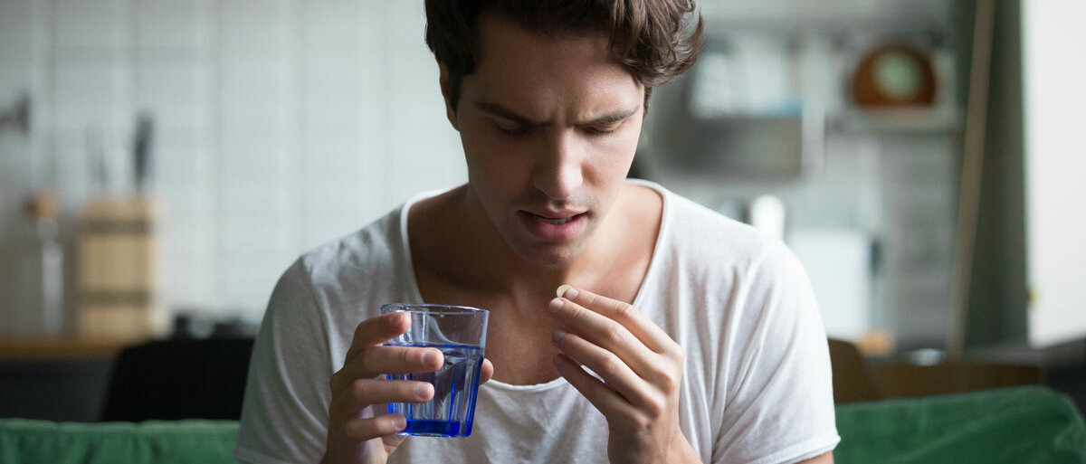 Ein junger Mann sitzt auf dem Sofa und verzieht das Gesicht. Er hält eine Tablette in der einen Hand, in der anderen ein Glas Wasser.