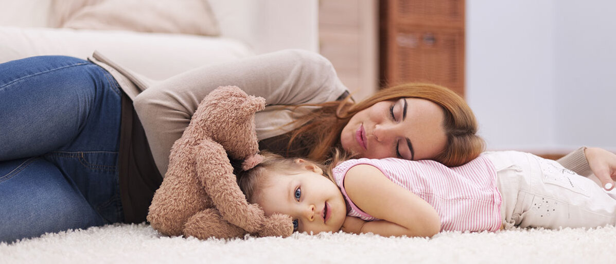 Mutter und Tochter liegen mit einem Teddy auf dem Teppich. Die Tochter ist hellwach, die Mutter ist auf dem Rücken der Tochter eingeschlafen.