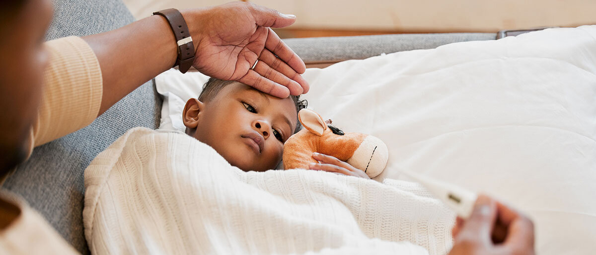 Frau legt bei ihrem Sohn die Hand auf die Stirn. In der anderen Hand hat sie ein Fieberthermometer. 
