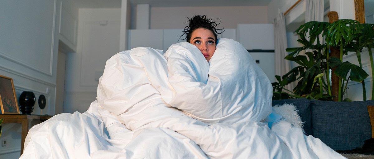 Eine Frau sitzt zu Hause auf dem Sofa und ist in eine große, weiße Bettdecke eingewickelt.