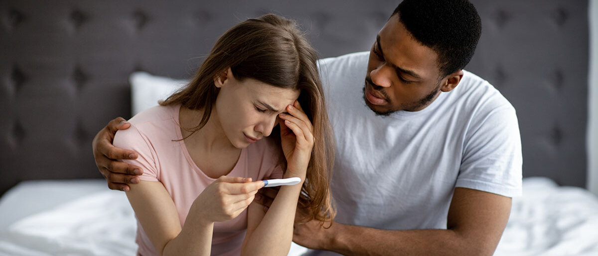 Ein junges Paar sitzt auf der Bettkante. Die Frau hält einen Schwangerschaftstest in der Hand. Die beiden halten sich traurig im Arm.