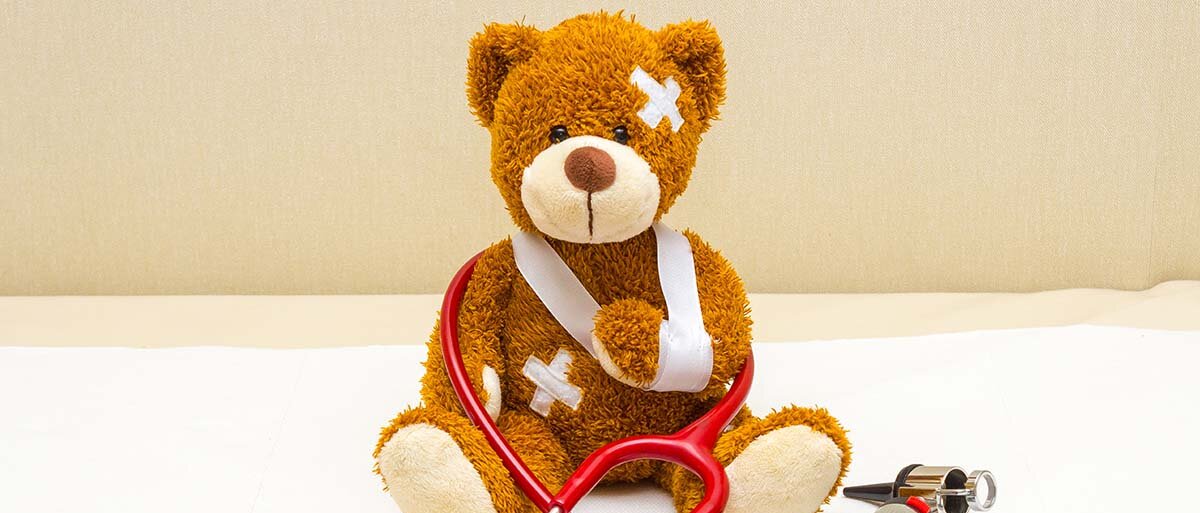 Teddybär mit Verbänden und Pflastern; um ihn gewickelt, ein rotes Stethoskop; neben ihm ein Otoskop