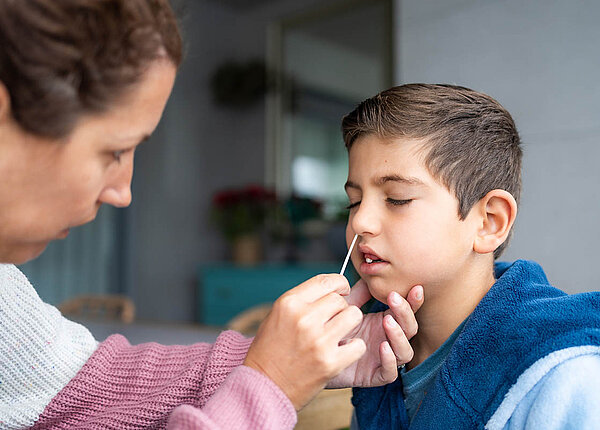 Eine Erwachsene steckt einem Jungen zu Hause ein Teststäbchen in die Nase.