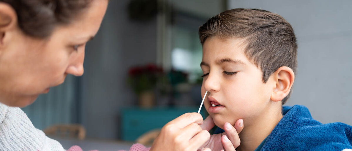 Eine Erwachsene steckt einem Jungen zu Hause ein Teststäbchen in die Nase.