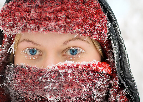 Eine Frau im Winter draußen hat ihr Gesicht mit einer Mütze und einem Schal gut verpackt. Mütze, Schal und Wimpern sind gefroren.