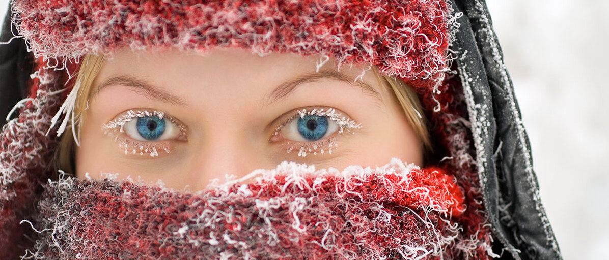 Eine Frau im Winter draußen hat ihr Gesicht mit einer Mütze und einem Schal gut verpackt. Mütze, Schal und Wimpern sind gefroren.