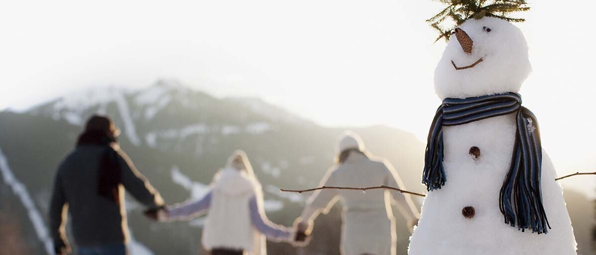 Im Vordergrund ein Schneemann; im Hintergrund die Rückansicht von drei Personen, die sich an den Händen halten und durch den Schnee stiefeln.