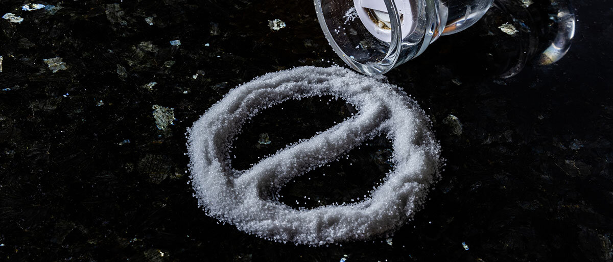 Auf einer schwarzen Tischplatte bilden Salzkristalle einen durchgestrichenen Kreis als Symbol für ein Verbot oder als Stoppzeichen. Daneben liegt der umgestürzte Salzstreuer.