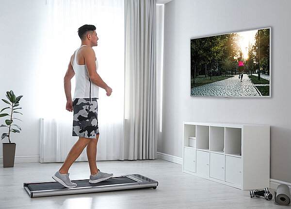Junger Mann laeuft auf einem Walking Pad vor einem Fernseher in einem Raum