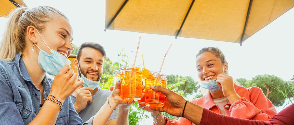 Vier junge Menschen sitzen unter einem Sonnenschirm am Tisch und prosten sich mit bunten Cocktails zu. Einige tragen Mund-Nasen-Schutz.