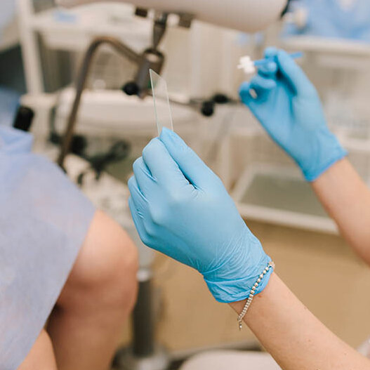 Eine Frau sitzt auf einem gynäkologischen Behandlungsstuhl. Die Frauenärztin hält in der linken Hand einen Objektträger für das Lichtmikroskop, in der rechten Hand ein Stäbchen, um einen Abstrich zu nehmen.