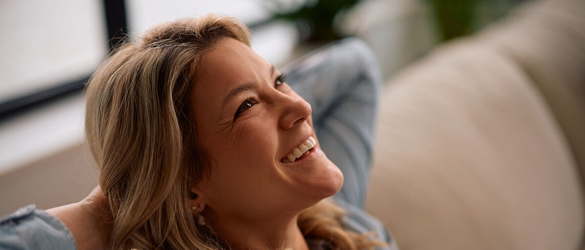 Wechseljahre bewusst gestalten Frau mittleren Alters sitzt glücklich auf der Couch, sie hat die Arme hinter dem Kopf verschränkt.