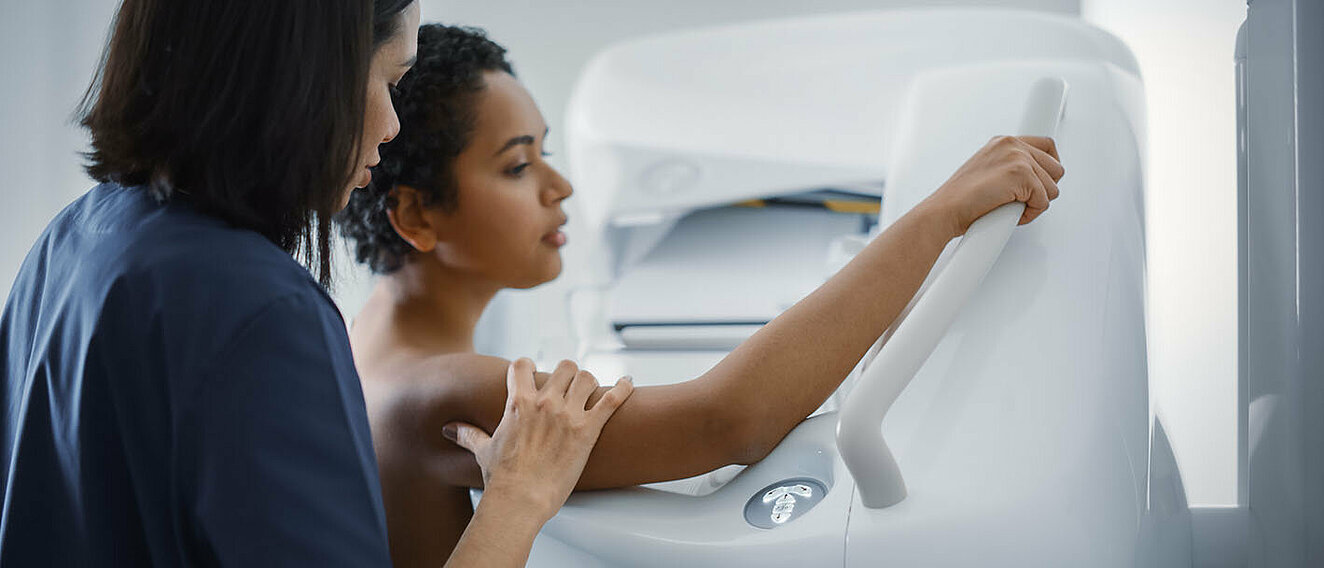Frau sitzt vor einem Mammographie Gerät. Die Ärztin steht neben ihr und unterstützt sie