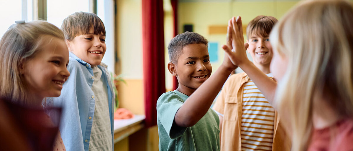 5 Schüler*innen in einem Klassenzimmer. Sie lachen. Zwei geben sich eine High-five. 