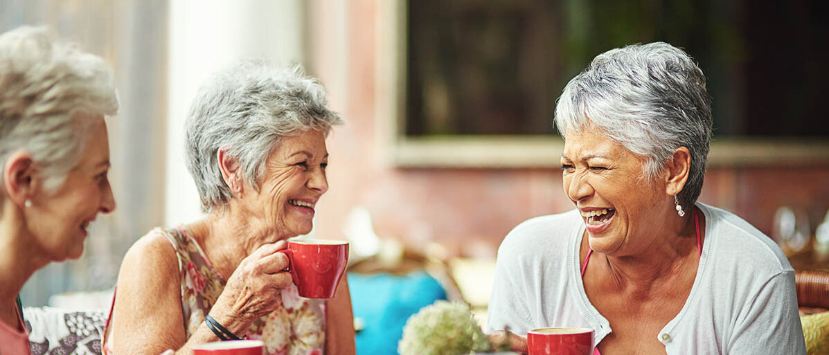 Kaffee und das Osteoporose-Risiko: so wirkt Koffein auf die Knochen von Frauen Drei Frauen sitzen im Freien an einem Tisch, jede hat eine rote Tasse vor sich. Zwei Frauen lachen.