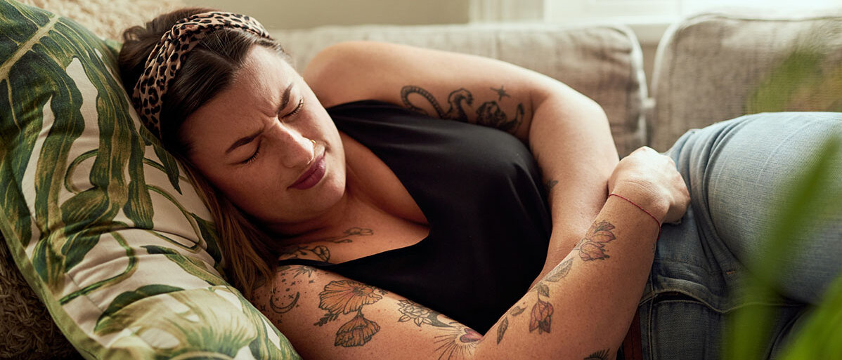 Eine Frau mit Tattoos an Armen und Dekolletee liegt auf dem Sofa. Sie hat die Augen geschlossen und das Gesicht verzerrt, die Beine angezogen und hält sich den Unterleib.