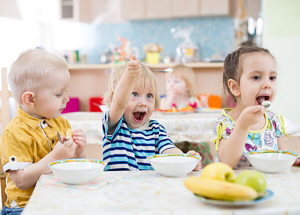 Drei Kinder sitzen in der Kita am Tisch und essen aus Schüsseln. Vor ihnen steht auch ein Teller mit Bananen und Äpfeln. Das mittlere Kind macht Quatsch mit seinem Löffel.