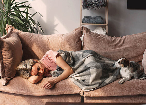 Eine Frau liegt traurig und in eine Decke gehüllt auf dem Sofa, ihr Hund liegt daneben.