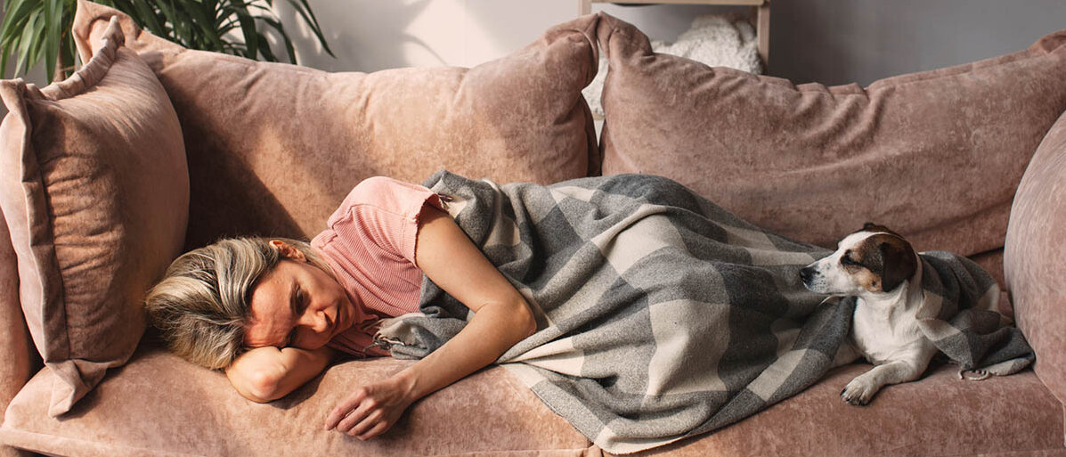 Eine Frau liegt traurig und in eine Decke gehüllt auf dem Sofa, ihr Hund liegt daneben.