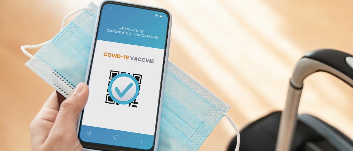 Eine Hand, die einen Reisepass, eine Maske und ein Handy in der Hand hält. Auf dem Bildschirm sieht man ein digitales COVID-19-Impfzertifikat.