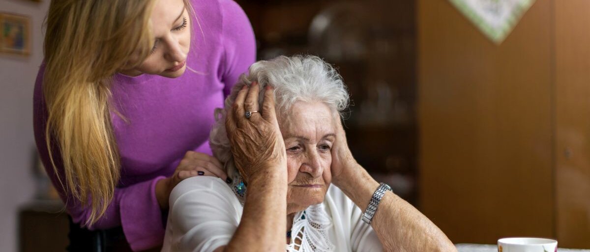 Eine junge Frau versucht einer Seniorin zu helfen, die an einem Tisch sitzt und sich an den Kopf fasst.