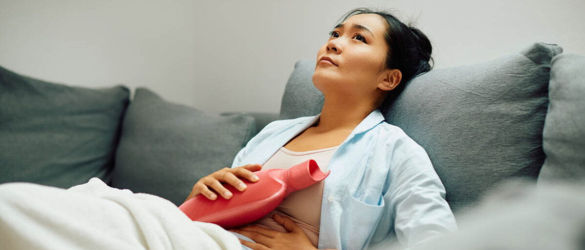 Menstruationsbeschwerden und Depression: Deshalb ist es wichtig, Periodenschmerzen zu behandeln. Eine junge Frau hat auf dem Sofa die Beine hochgelegt und hält sich eine Wärmflasche auf den Bauch.