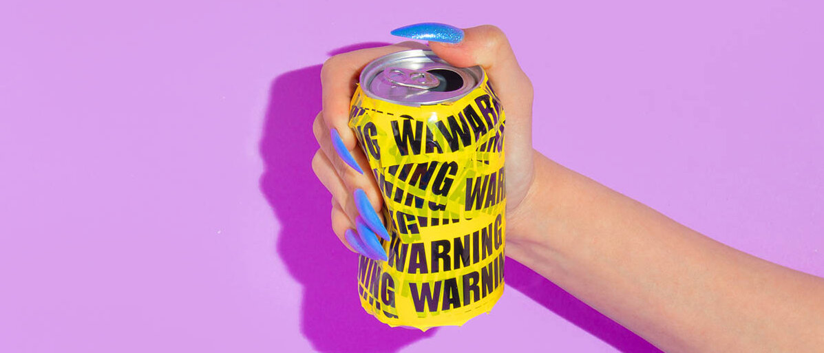 Eine Frau zerdrückt eine Getränkedose in der Hand. Die Dose ist mit gelbem Flatterband umwickelt, auf dem vielfach „WARNING“ steht.