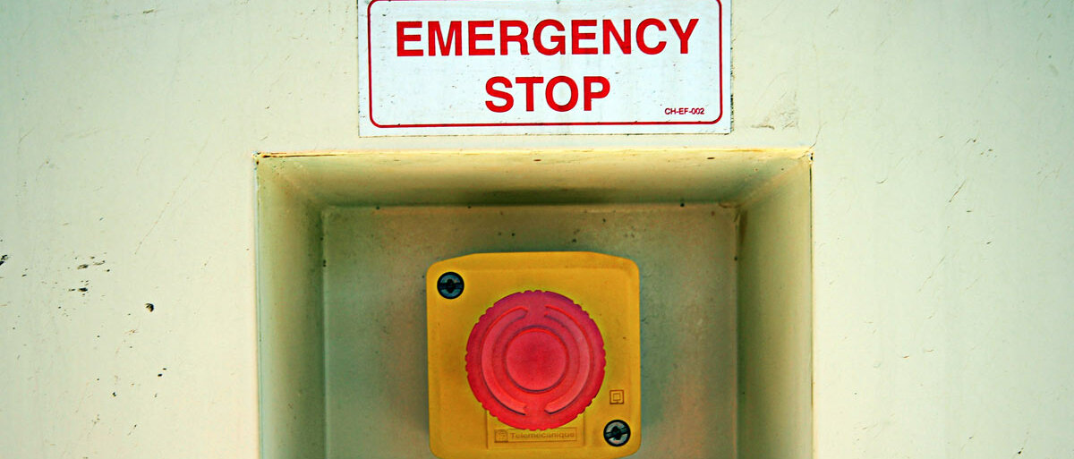 Ein rot-gelber Notaus-Schalter hängt an der Wand, darüber ein Schild mit der Aufschrift "Emergency Stop".