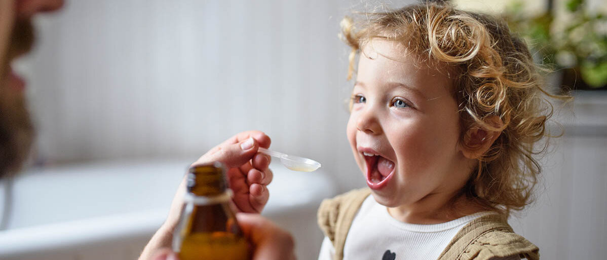 Suspensionen herstellen in der Rezeptur: Praxiswissen und häufige Fehler Ein Mann gibt einem Kleinkind mit einem Dosierlöffel eine Suspension, es hat den Mund weit offen. In der anderen Hand hält der Mann die Braunglasflasche.