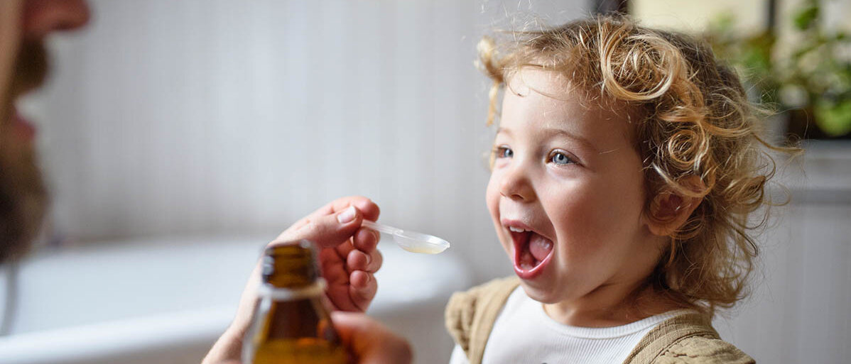 Ein Mann gibt einem Kleinkind mit einem Dosierlöffel eine Suspension, es hat den Mund weit offen. In der anderen Hand hält der Mann die Braunglasflasche.
