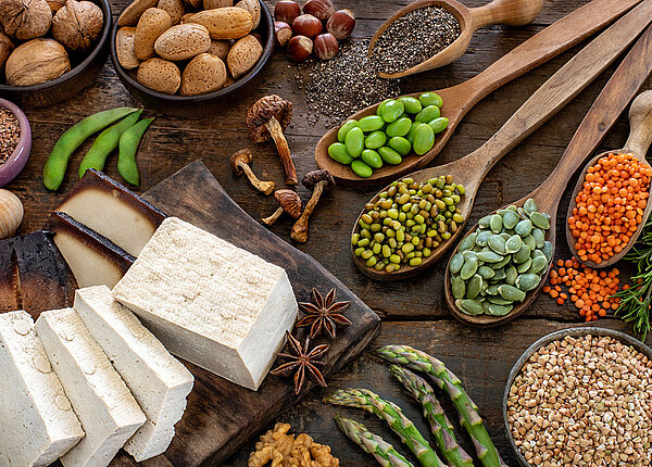 In Scheiben geschnittener Tofu auf einem Schneidebrett, daneben verschiedene Hülsenfrüchte wie Linsen, Edamame und Erbsen jeweils auf einem Holzlöffel
