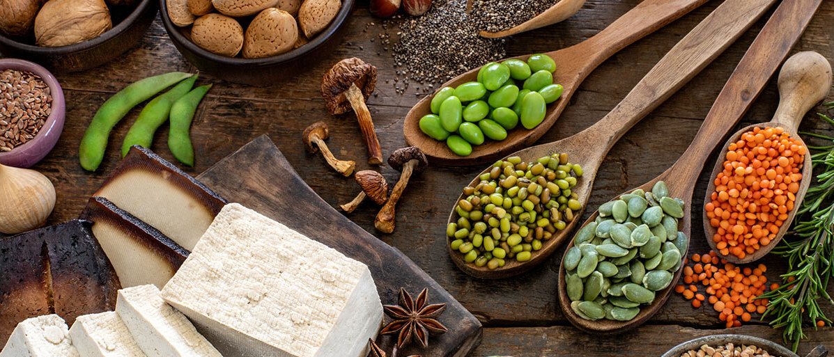 In Scheiben geschnittener Tofu auf einem Schneidebrett, daneben verschiedene Hülsenfrüchte wie Linsen, Edamame und Erbsen jeweils auf einem Holzlöffel