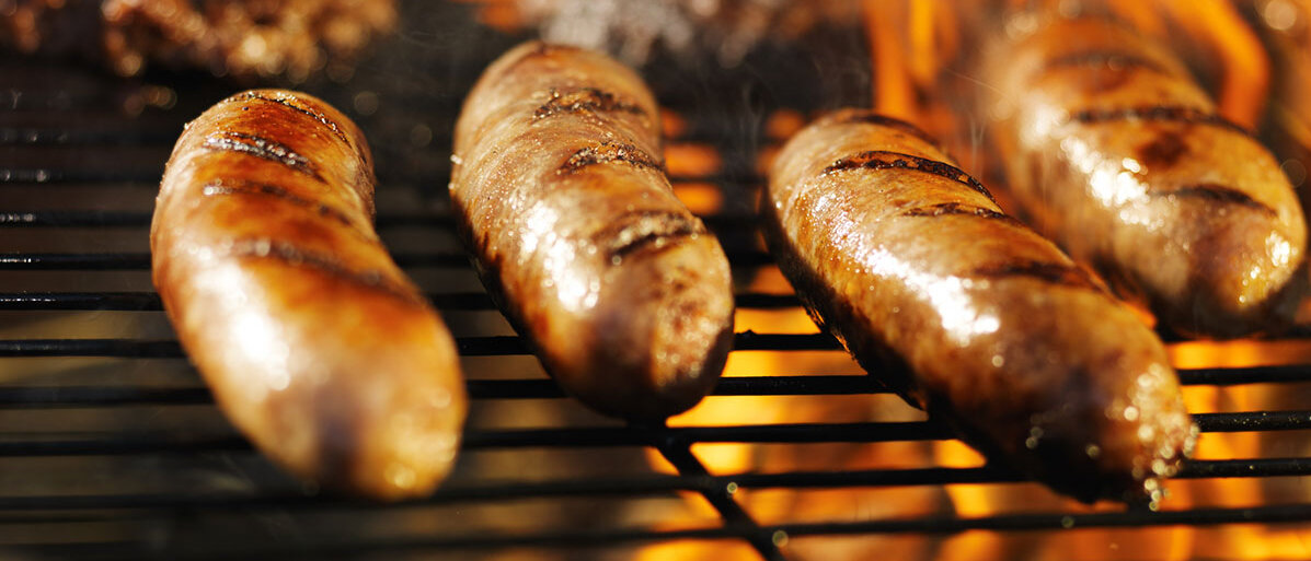 Im Vordergrund vier Würstchen auf einem Grillrost, im Hintergrund ein paar Steaks mit Flammen