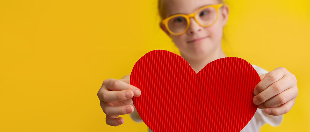 Ein Mädchen mit Down-Syndrom vor einem gelben Hintergrund: Das Mädchen trägt eine gelbe Brille, lächelt und hält dem Betrachter ein Herz aus roter Wellpappe entgegen.