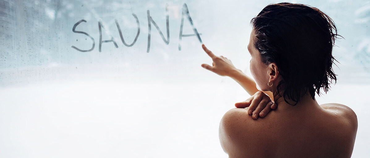 Sauna Frau von hinten in Saunahandtuch gewickelt. Sie schreibt mit ihrem Finger „Sauna“ auf eine beschlagene Scheibe.