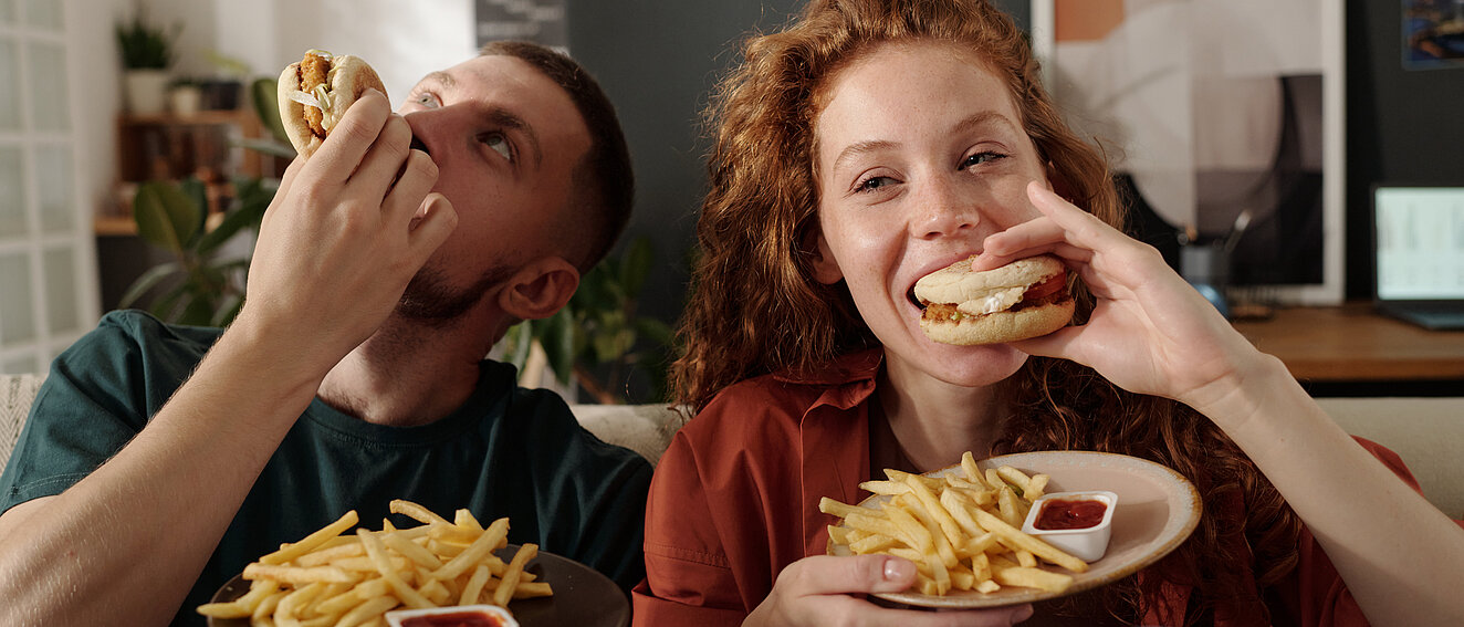 eine Frau und ein Mann sitzen nebeneinander uns essen jeder einen Burger mit einer Portion Pommes und Ketchup. Sie sehen sehr glücklich aus. 