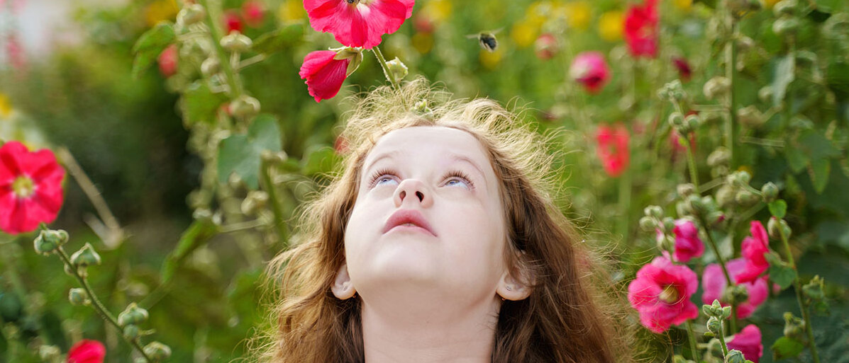 Kleines Mädchen umringt von Blumen schaut auf zu einer fliegenden Biene.