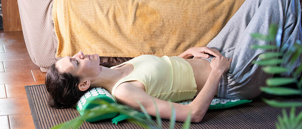 Akupressurmatte im Check: Hilft Akupressur bei RĂŒckenschmerzen, zur Muskelentspannung und Durchblutung? Eine Frau liegt auf dem RĂŒcken auf einer Akupressurmatte auf dem Boden vor ihrem Sofa. Sie hat entspannt die Augen geschlossen.