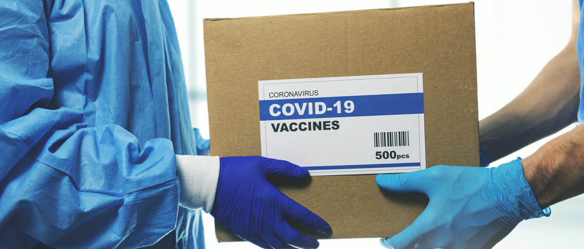 Eine Person in Schutzausrüstung übergibt einer anderen einen Karton mit COVID-19-Impfstoffen.