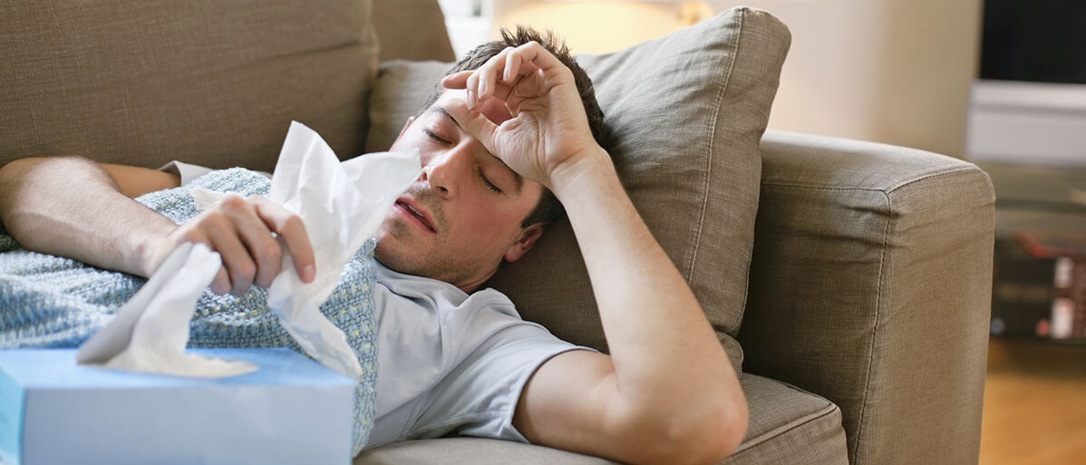 Mann liegt krank auf der Couch und haelt Taschentuch in der Hand. Eine Hand hat er am Kopf