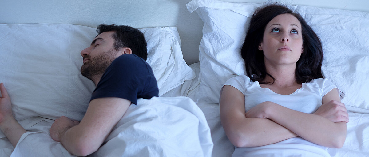 Ein Paar liegt im Bett, die Frau schaut wach zur Decke, der Mann liegt auf der Seite und schläft