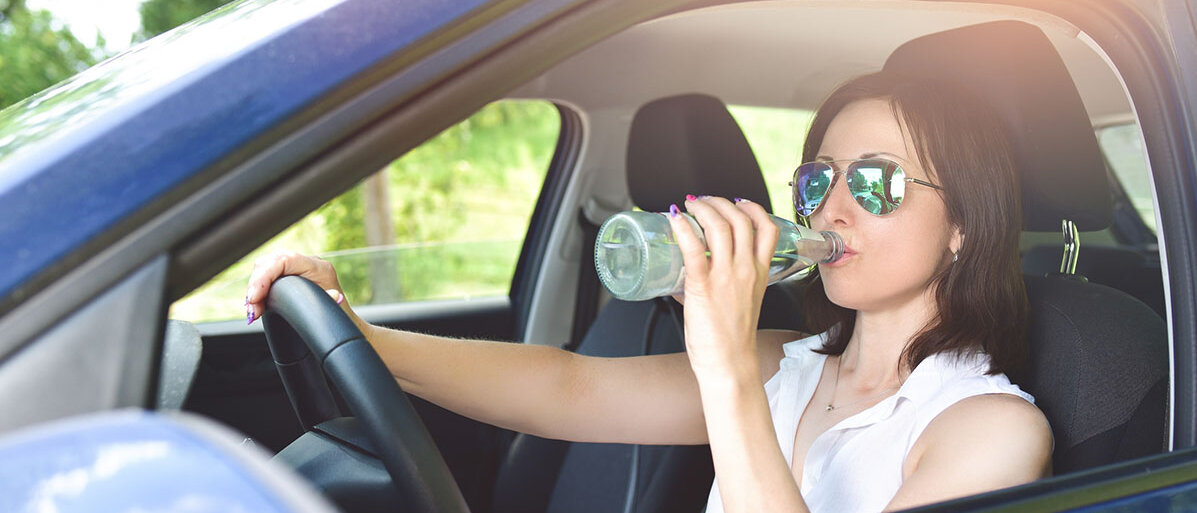 Frau mit Sonnenbrille sitzt am Steuer im Auto und trinkt Wasser aus einer Flasche